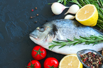 Fresh raw dorado fish with rosemary, garlic, tomatoes, pepper and lemon on a black slate background. Selective focus. Overhead view. Copy space.