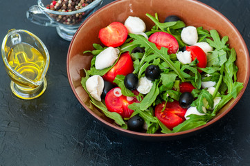Close up of fresh salad with arugula, tomatoes, olives, onion, mozzarella and spices in a bowl on a gray slate background. Healthy food, Diet, Detox, Clean Eating or Vegetarian concept