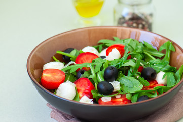 Close up of fresh salad with arugula, tomatoes, olives, onion, mozzarella and spices in a bowl on a white stone background. Healthy food, Diet, Detox, Clean Eating or Vegetarian concept