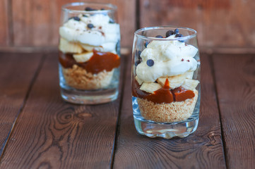 Dessert in a glass - Banoffee pie from bananas, crumble, salted caramel and wipped double cream and chocolate chips on a wooden background. Close up and copy space.