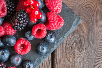 Mix of berries raspberries red currants blueberries and blackberries on black slate board. Wooden background.  Overhead view and copy space