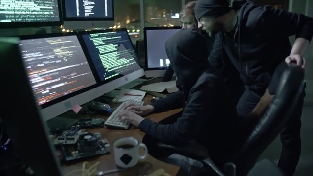 Tilt up of team of hackers typing programming code on computer monitors in office at night