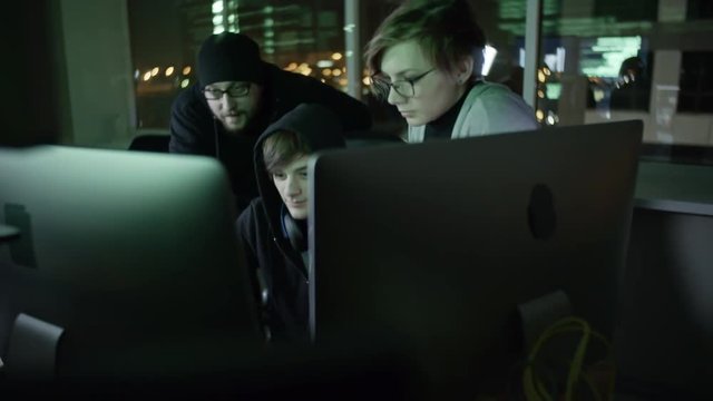 Tilt down of three cybercriminals working in team in front of computers in the dark office