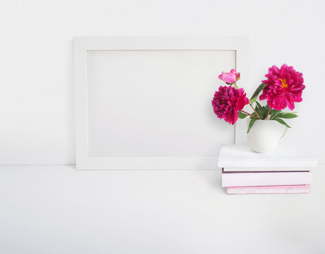 White blank wooden frame mockup with a peony flowers bouquet in a porcelain cup and pile of books lying on the table. Poster product design. Styled stock feminine photography. Home decor.