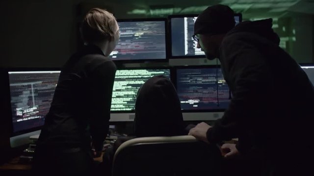 Rear view of three computer programmers at desk in front of several monitors and writing codes