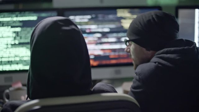 Tracking shot rear view of three hackers discussing how to crack network and typing on computers at night