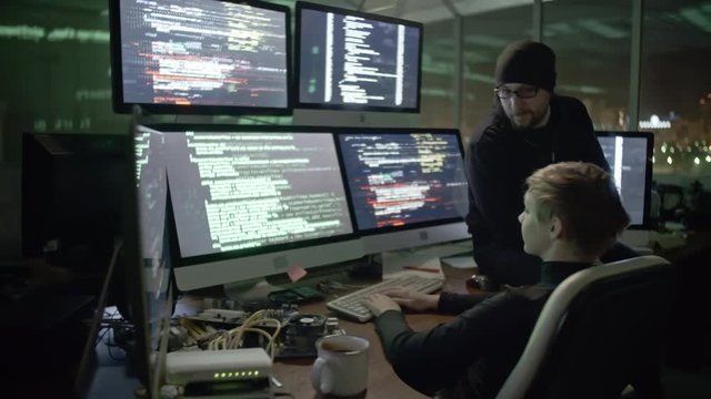 Young woman typing program codes on computer screens while cracking website with male colleague. Man sitting on table and telling her something