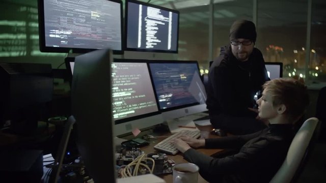 Female hacker working with data on computer screens in dark room with male colleague. Green codes reflected on walls