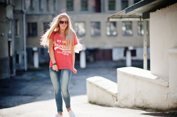 Stylish happy blonde woman wear at jeans, sunglasses and t-shirt posed at street on sunny weather. Fashion urban model portrait.