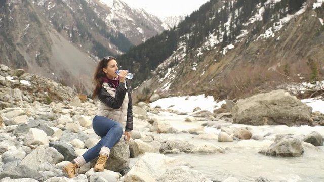 beautiful woman traveler drinks water near a mountain river on a background of mountains