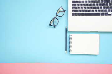 Flat lay design of work desk with labtop, notebook, glasses and pencil on pink and blue background.
