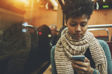 Pensive afro american student travelling home after study in evening and messaging with family via smartphone, serious black teenage female with curly hair in train coach using her cell telephone