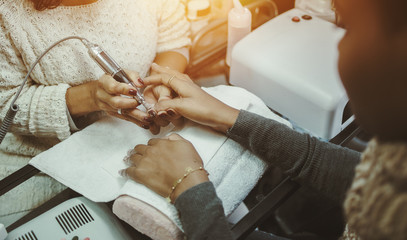 Close-up view of hands of afro american brown woman in nail salon receiving manicure, master preparing nails for shellac manicure using special rotating nail file machine device