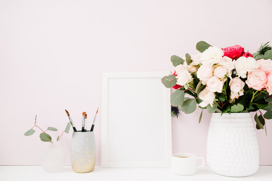 Home office desk with photo frame mock up, beautiful roses and eucalyptus bouquet in front of pale pastel pink background. Blog, website or social media concept .