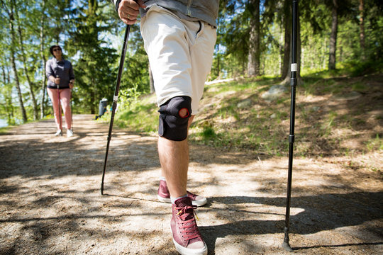 Man with bandage on knee. Rehabilitative sport in Finland - nordic walking. Man and mature woman hiking in green sunny forest. Active people outdoors. Scenic peaceful Finnish summer landscape.