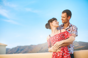 happiness couple stands on the veranda under sky on mountain background. smiling man in shirt hugs a woman in red dress