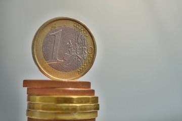Side view of tall pile of metal coins in golden and copper color in front of silver background with one Euro (European currency EUR) coin on the top 