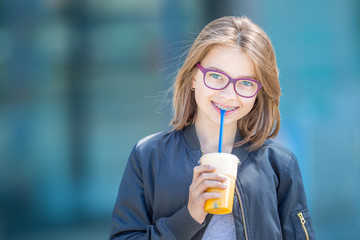 Cheerful pre-teenage girl drinking juice. Girl with dental teeth braces and glasses.