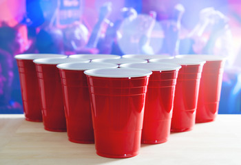 Beer pong tournament layout. Many red party cups in a nightclub full of people dancing on the dance floor in the background. Perfect for marketing and promotion for event. Plastic mugs on wooden table