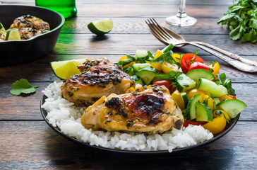 Spicy and crispy homemade cilantro lime chicken thighs in a black cast-iron skillet and on a plate with vegetable salad and white rice. Wooden rustic background.