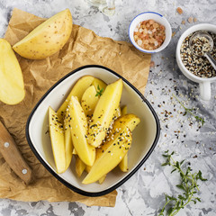 Slices of raw potatoes in seeds of dark and white sesame seeds in a white enamel bowl before baking in the oven. Top view on a marble gray background