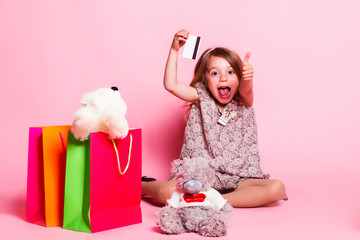 Little girl Shows thumb and tongue on a pink background with a teddy bear, shopping bags