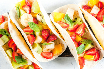 Summer snacks. Food for a party. Fruit tacos with strawberries, mangoes, bananas, chocolate, mint. On a light blue concrete table. Copy space