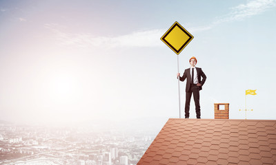 Fototapeta premium Young businessman on house brick roof holding yellow signboard and looking at city. Mixed media