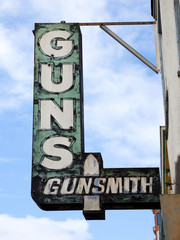 Weathered GUNS GUNSMITH sign attached to building against blue sky. Vertical.