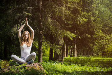 Girl doing yoga in nature. Young beautiful blonde, sits on a stump in nature in a Burmese pose and...