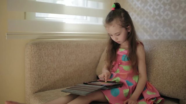 Little cute girl sitting on sofa and playing with tablet