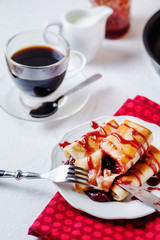 Hearty, tasty and traditional breakfast or lunch, in a hotel or restaurant, fried thin pancakes with strawberry or cherry jam, syrup, black coffee and milk on a light background