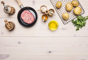 cooking homemade hamburger with fried potatoes, fresh ground beef in a small pan with parsley, different spices on white wooden table border place for text  top view close up