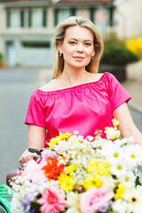 Obraz na płótnie Canvas Outdoor close up portrait of blond woman wearing bright pink dress, holding bicycle with big bouquet of flowers in basket