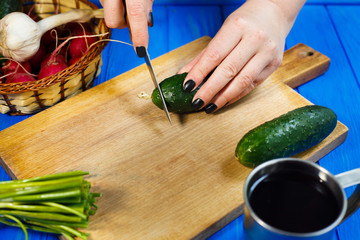 Woman hands cutting fresh crunchy cucumber on cutting board with spring onions and herbs on the foreground