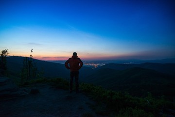 Man tourist standing on mountaintop observing city lights.