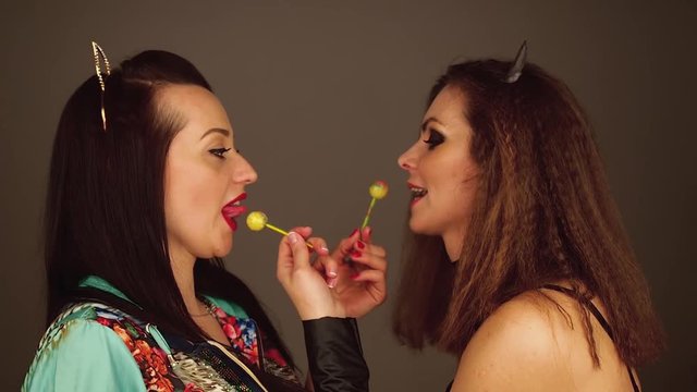 Two young women suck on lollypops looking at each other and lauphing.