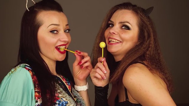 Two young women suck on lollypops looking at each other and lauphing.