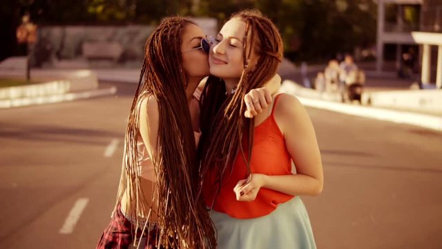 Two happy women with dreads walking on the empty road and talking in summer. Two hipster girls laughing, hugging and kissing each other during a bright sunny day. Slowmotion shot.