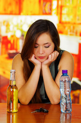 Portrait of a beautiful drunk woman posing her hands in her chin, with a bottle of beer and water and car keys over a wooden table, in bar background