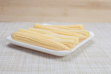 baby corn on a white plate on a brown cloth over wooden background