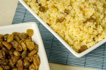 Plate with freshly cooked Moroccan couscous and plate with with raisins on blue straw background