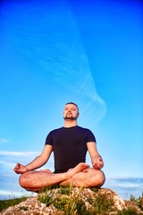 Portrait of the man sitting on a rock in the lotus position against blue sky.