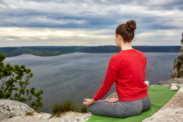 Naklejka na ściany i meble Young woman meditating in a lotus pose on the rock above big river.