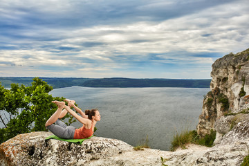 Young sportive woman training yoga on rock against beautiful big river.