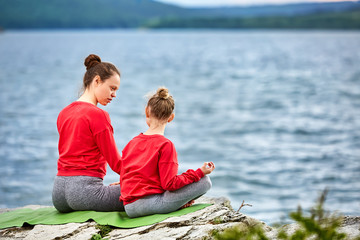 Rear view of the mother and daughter are practicing yoga on the rock near river.