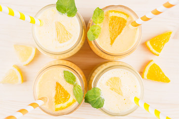 Freshly blended orange and yellow lemon smoothie in glass jars with straw, mint leaf, top view, close up. White wooden board background.