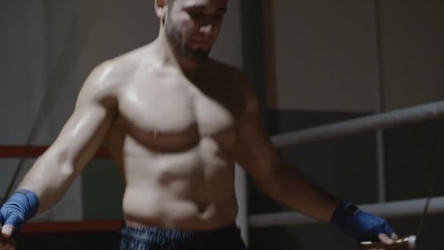 Tilt down of shirtless muscular athlete skipping rope in boxing ring of dark gym in slow motion