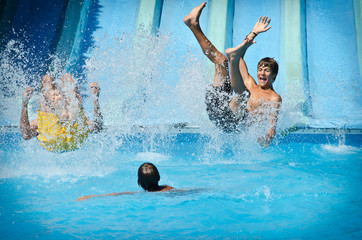 Young people having fun on water slides in aqua park, splashing into swimming pool