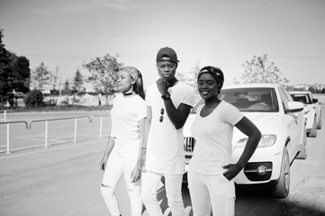 Three stylish african american friends, wear on white clothes against two luxury cars. Street fashion of young black people. Black man with two african girls.